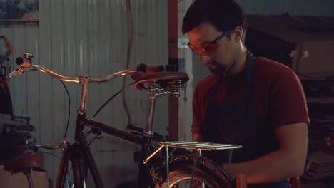 theme-small-business-bike-repair.-A-young-Caucasian-brunette-man-wearing-safety-goggles,-gloves-and-an-apron-uses-a-hand-tool-to-repair-and-adjust-the-bike-in-the-workshop-garage