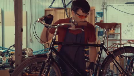 theme-small-business-bike-repair.-A-young-Caucasian-brunette-man-wearing-safety-goggles,-gloves-and-an-apron-uses-a-hand-tool-to-repair-and-adjust-the-bike-in-the-workshop-garage