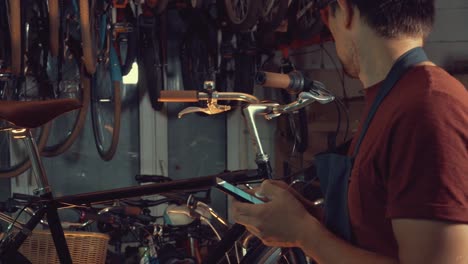 theme-small-business-bike-repair.-Young-caucasian-brunette-man-wearing-safety-goggles,-gloves-and-fartukhe-uses-mobile-phone-technology,-takes-notes,-checklist-in-bicycle-workshop