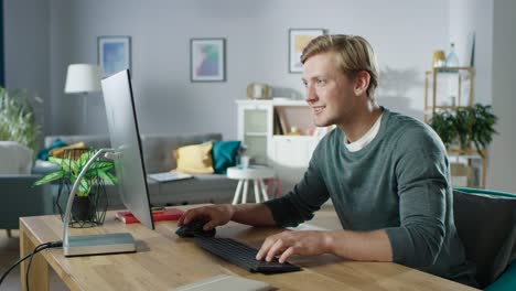 Portrait-of-the-Focused-Young-Man-Working-on-a-Personal-Computer-while-Sitting-at-His-Desk.-In-the-Background-Cozy-Living-Room.