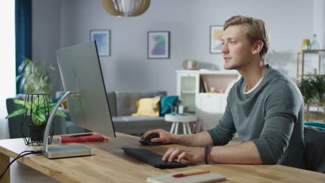 Portrait-of-the-Handsome-Man-Working-on-Personal-Computer-while-Sitting-at-His-Desk.-In-the-Background-Stylish-Cozy-Living-Room.-Young-Freelancer-Working-From-Home.