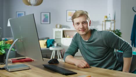 Portrait-of-the-Handsome-Young-Man-Looking-at-the-Camera-While-Sitting-at-His-Desk-with-Personal-Computer.-In-the-Background-Cozy-Living-Room.