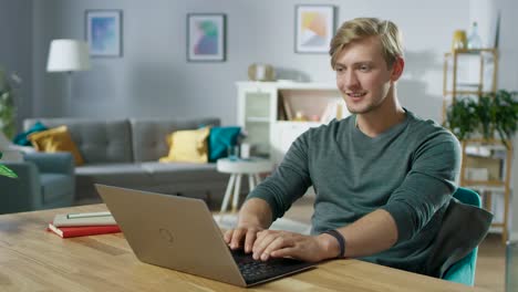 Guapo-sonriente-joven-usando-el-portátil-mientras-está-sentado-en-el-escritorio-de-su-acogedora-sala-de-estar.