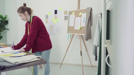 Junge-asiatische-Frau-Mode-Designer-Zeichnung-mit-Bleistift-und-Papier-während-der-Arbeit-im-Workshop-Studio-betrachten.