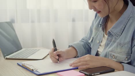 Beautiful-smart-Asian-young-entrepreneur-business-woman-owner-of-SME-checking-product-on-stock-and-write-on-clipboard-working-at-home.-Small-business-owner-at-home-office-concept.