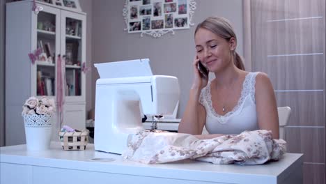 Mujer-joven-felizmente-hablando-por-teléfono-mientras-está-sentado-en-la-mesa-junto-a-la-máquina-de-coser