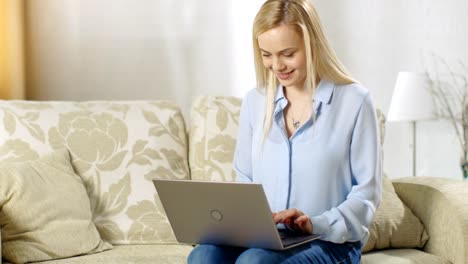 In-the-Living-Room.-Young-Blonde-Woman-Sitting-on-a-Sofa-Types-on-a-Laptop-that-is-Situated-on-Her-Laps.