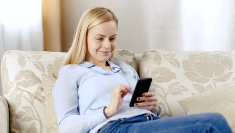 Beautiful-Young-Blonde-Sitting-in-Her-Living-Room-on-a-Sofa.-Smiling,-She-Uses-Smartphone.