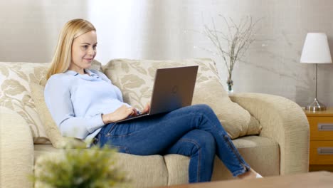 Beautiful-Blonde-Woman-Sitting-on-a-Couch-with-Notebook-on--Her-Lap,-She-Actively-Types-on-It.-Her-Room-is-Full-of-Light.