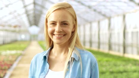 En-un-día-soleado-hermoso-jardinero-rubia-está-sonriendo-en-un-invernadero-lleno-de-flores-coloridas.