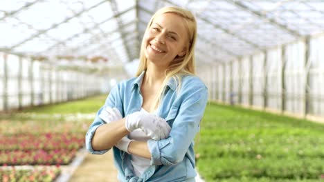 On-a-Sunny-Day-Beautiful-Blonde-Gardener-Stands-Smiling-in-a-Greenhouse-Full-of-Colorful-Flowers.