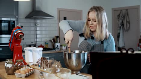 Young-beautiful-woman-mixing-the-ingredients-in-a-bowl,-preparing-dough-for-baking.-Blonde-female-in-kitchen