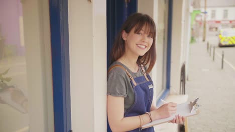 Confident-Young-Small-Business-Owner-Writing-In-Clipboard-Outside-Store