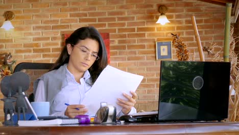 Dolly-shot-of-Hispanic-young-woman-working-at-home-office,-reading-documments-and-working-on-laptop-computer.