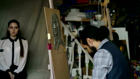 Sculptor-creating-sculpture-of-human's-face-on-canvas-while-young-woman-posing-to-him-in-art-studio