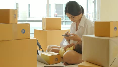Young-asian-mother-holding-baby-and-checking-order-from-smartphone-for-customer-and-online-delivery-for-ready-packing-in-bedroom.