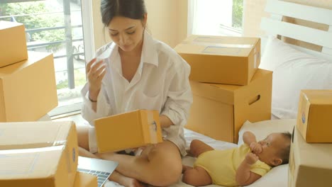 Young-asian-mother-and-her-newborn-child-checking-order-from-laptop-for-customer-and-online-delivery-for-ready-packing-in-bedroom.