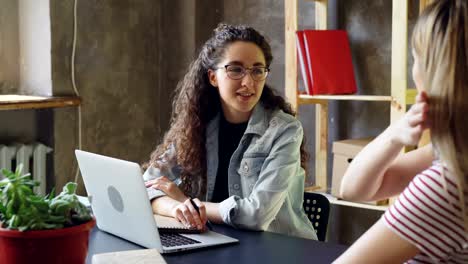 Joven-dueño-de-la-pequeña-empresa-es-hablar-con-su-cliente-en-la-oficina-moderna.-Las-mujeres-se-sientan-en-el-escritorio,-charlando-y-riendo-juntos-de-manera-amistosa.