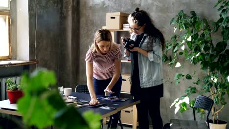 Young-female-enterpreneurs-are-placing-photos-on-table-to-make-flat-lay-and-shooting-with-camera-while-standing-in-modern-office.-Women-are-sharing-ideas-and-discussing-design.