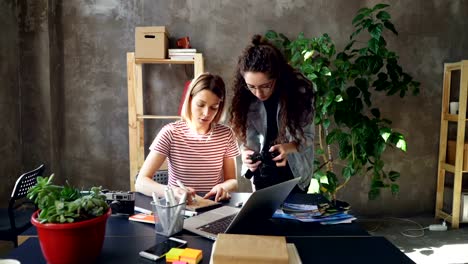 Young-designer-is-drawing-images-in-notebook-sitting-at-table-while-female-photographer-is-coming-to-her-with-camera.-Women-start-discussing-project-and-watching-photos-on-screen.