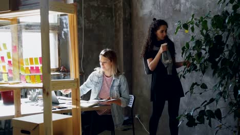 Female-friends-and-business-partners-are-working-together-in-modern-office.-Blonde-is-sitting-at-table-and-collecting-pictures,-brunette-is-watering-plants.