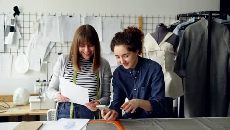 Creative-dressmakers-are-using-smartphone-and-looking-at-clothing-sketches-while-working-in-nice-studio.-Dummy,-sewing-machine-and-women's-garments-in-background.