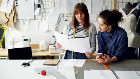 Young-women-working-in-design-industry-are-comparing-sketches-and-talking-about-them-while-working-in-modern-tailor-shop.-Productive-teamwork-concept.