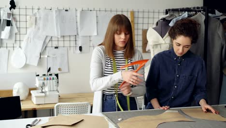 Equipo-de-diseñadores-creativos-trabaja-con-patrones-de-ropa-y-tela-en-sastrería.-Las-mujeres-jóvenes-se-concentran-en-la-medición.-Artículos-de-coser-y-herramientas-de-fondo.