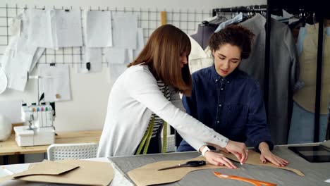 Experienced-seamstress-is-teaching-her-assistant-to-outline-clothing-patterns-on-fabric.-Young-woman-is-focused-on-process,-watching-carefully-and-asking-questions.