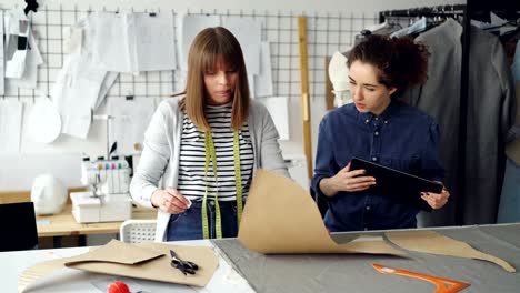 Young-tailors-are-working-with-tablet,-communicating-and-outlining-clothing-pattern-on-textile-on-studio-desk.-Light-workshop-with-sewing-items-in-background.