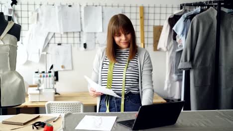 Young-attractive-woman-is-choosing-sketches-for-new-fashion-show-and-using-laptop-in-her-tailor's-shop.-Creative-thinking-in-clothing-design-industry-concept.