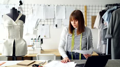 Diseñador-de-ropa-profesional-es-mirando-a-través-de-dibujos,-añadir-pequeños-detalles-y-comprobar-portátil-en-su-taller-de-luz.-Tecnologías-modernas-para-el-concepto-de-inspiración.