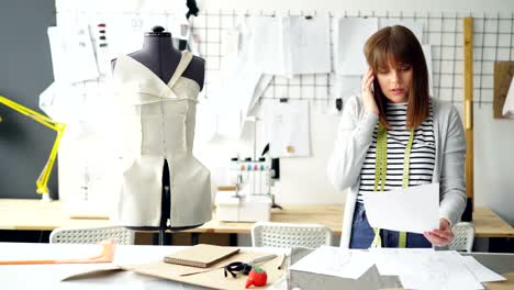 Self-employed-seamstress-is-talking-on-mobile-phone-and-looking-through-clothing-sketches-in-her-studio.-Sewing-items,-dummy-and-drawings-pinned-on-wall-are-visible.