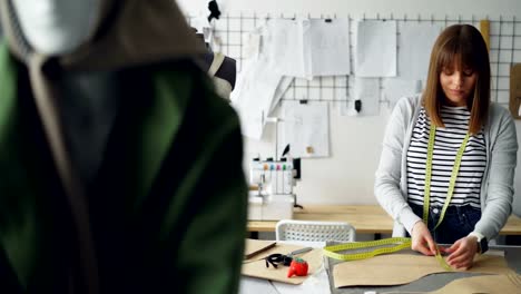 Attractive-female-tailor-is-measuring-clothing-patterns-with-tape-measure-in-her-light-tailor's-shop.-Everyday-work-in-clothes-manufacturing-industry-concept.