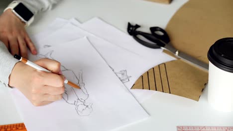 Close-up-shot-of-woman's-hand-drawing-outlines-of-stylish-ladies'-garment.-Paper,-tailor's-scissors,-ruler-and-coffee-are-visible-on-studio-table.-Designer-at-work-concept.