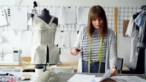 Young-ambitious-seamstress-is-taking-photos-of-garment-sketches-lying-on-table-in-her-modern-light-studio-using-smartphone.-Woman-is-confident-and-concentrated.