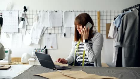 Small-business-owner-is-ordering-fabric-for-tailor's-shop-with-mobile-phone-and-working-with-laptop.-Woman-is-talking-and-typing-simultaneously.-Modern-means-of-communication-concept.