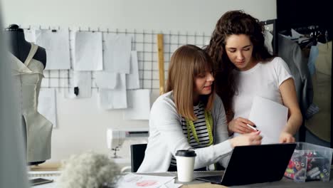 Zwei-attraktive-Näherinnen-sind-mit-Blick-auf-Laptop-und-Nähfäden-für-Kleidungsstück-auf-dem-Bildschirm-auswählen.-Erstellen-neue-Kleider,-die-mit-moderner-Technik-Konzept.