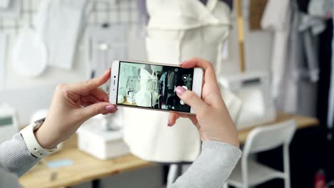 Close-up-shot-of-women's-hands-holding-smartphone-and-photographing-tailoring-dummy-with-half-finished-garment-pinned-to-it.-Modern-technologies-in-small-business-concept.