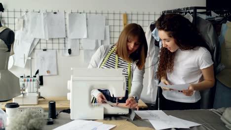 Junge-Frau-arbeitet-mit-Nähmaschine-und-Stiche-überprüfen,-wenn-ihre-Kollegin-mit-Skizze-zu-ihr-kommt.-Frauen-sind-mit-Blick-auf-Stoff-und-sprechen.