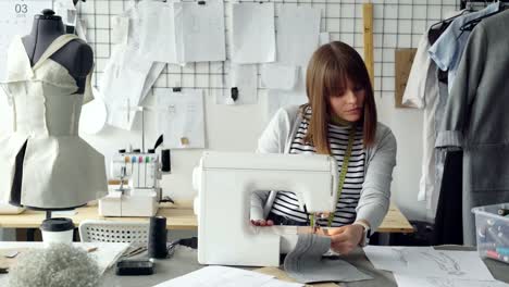 Junge-attraktive-Schneiderin-arbeitet-an-der-Nähmaschine-und-mit-Blick-auf-Frauen-Kleidungsstück-Skizzen-in-ihrem-Atelier-am-Tisch.-Moderne-Ausstattung-und-modische-Kleidung-im-Hintergrund.