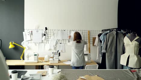 Back-view-of-creative-female-tailor-taking-sketches-from-tailoring-desk-and-hanging-them-on-wall-above-table.-Planning-and-creating-new-collection-concept.