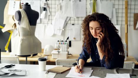 Professional-fashion-designer-is-talking-on-mobile-phone-and-drawing-clothing-sketch-with-pencil-at-table-in-tailoring-shop.-Everyday-work-and-communication-in-workplace-concept.