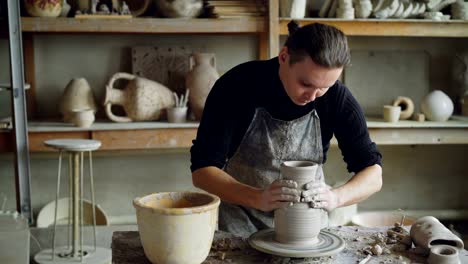 Handsome-young-man-is-molding-ceramic-vase-from-clay-on-spinning-throwing-wheel-while-working-in-potter's-studio.-Professional-is-concentrated-on-work.