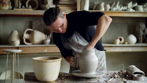 Skillful-young-potter-is-molding-ceramic-vase-from-clay-on-turning-throwing-wheel-while-working-in-potter's-workshop.-Professional-is-concentrated-on-work.