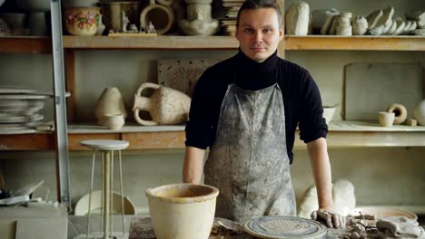 Porträt-von-hübschen-jungen-Potter-in-schlammigen-Schürze-stehen-am-Tisch-in-Werkstatt-und-Blick-in-die-Kamera.-Regale-mit-handgefertigten-Vasen-und-Töpfe-im-Hintergrund.