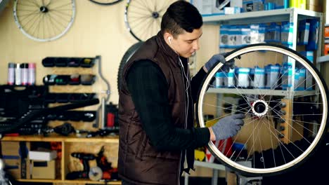 Young-serviceman-is-cleaning-bicycle-wheel-outside-and-inside-with-tools-and-piece-of-cloth-and-listening-to-music-with-earphones.-Profession-and-people-concept.