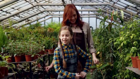 Joven-jardinero-mujer-en-delantal-y-su-linda-hija-bailan-en-invernadero-que-se-divierten.-Concepto-de-familia,-jardinería,-padres-e-hijos-felices.