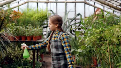 Chica-guapa-es-caminar-en-invernadero-y-rociando-agua-sobre-la-vegetación-mientras-que-su-madre-ocupada-trabaja-en-segundo-plano.-Infancia,-ayudando-a-los-padres-y-el-concepto-de-familia.