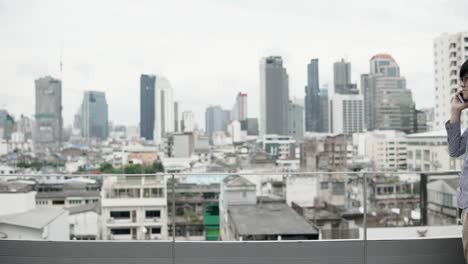 Young-Asian-businessman-using-smartphone-for-business-talk-walking-on-office-building-rooftop-terrace-with-city-view-in-the-background.-Business-communication-and-corporation-concepts.